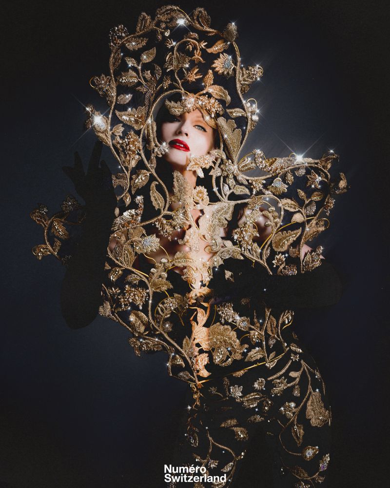 A woman in ornate gold floral gown and headdress with red lipstick poses in a dramatic studio shot for Numero Switzerland.