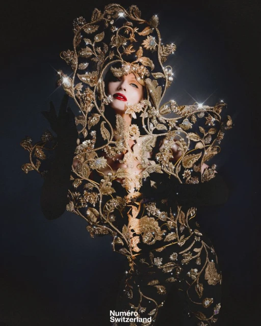 A woman in ornate gold floral gown and headdress with red lipstick poses in a dramatic studio shot for Numero Switzerland.