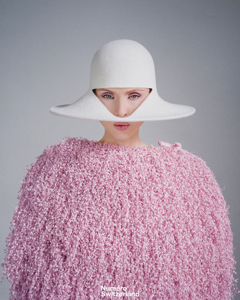 Close-up fashion portrait of a woman wearing a large white hat and pink sweater, set against a neutral background. Beauty shot.