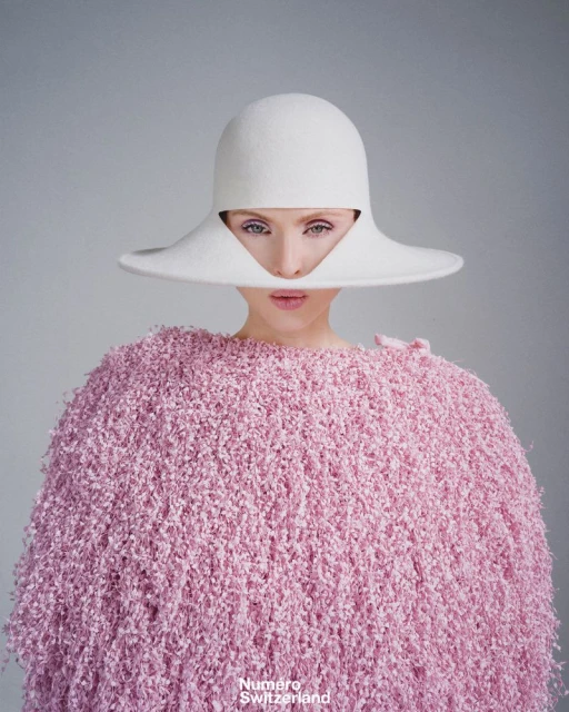 Close-up fashion portrait of a woman wearing a large white hat and pink sweater, set against a neutral background. Beauty shot.