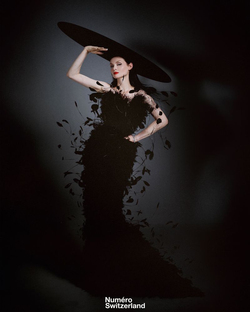 Elegant woman in a feathered gown and wide-brimmed hat posing in a studio against a dark grey background for a fashion editorial.