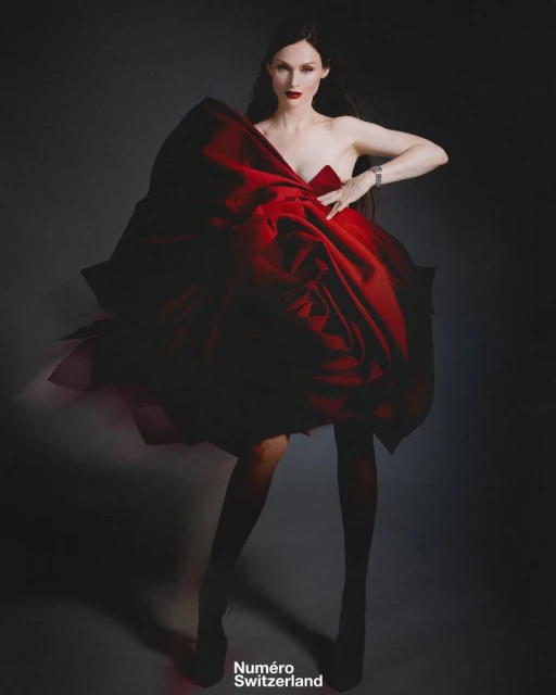 Elegant fashion shot of a woman in a large red dress and wristwatch posing against a dark backdrop in a studio with mood lighting.