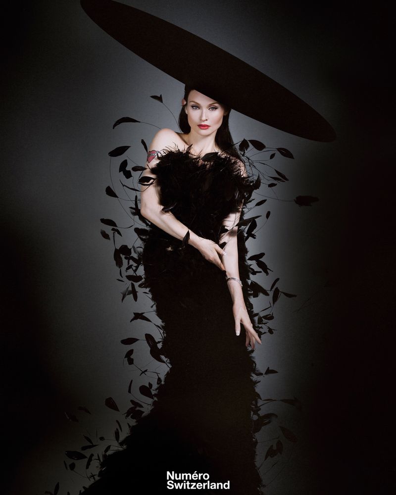 A woman poses in a black dress with feathers and a large hat against a gradient grey background in a studio fashion portrait.