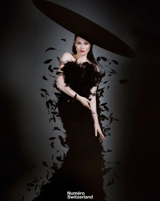 A woman poses in a black dress with feathers and a large hat against a gradient grey background in a studio fashion portrait.