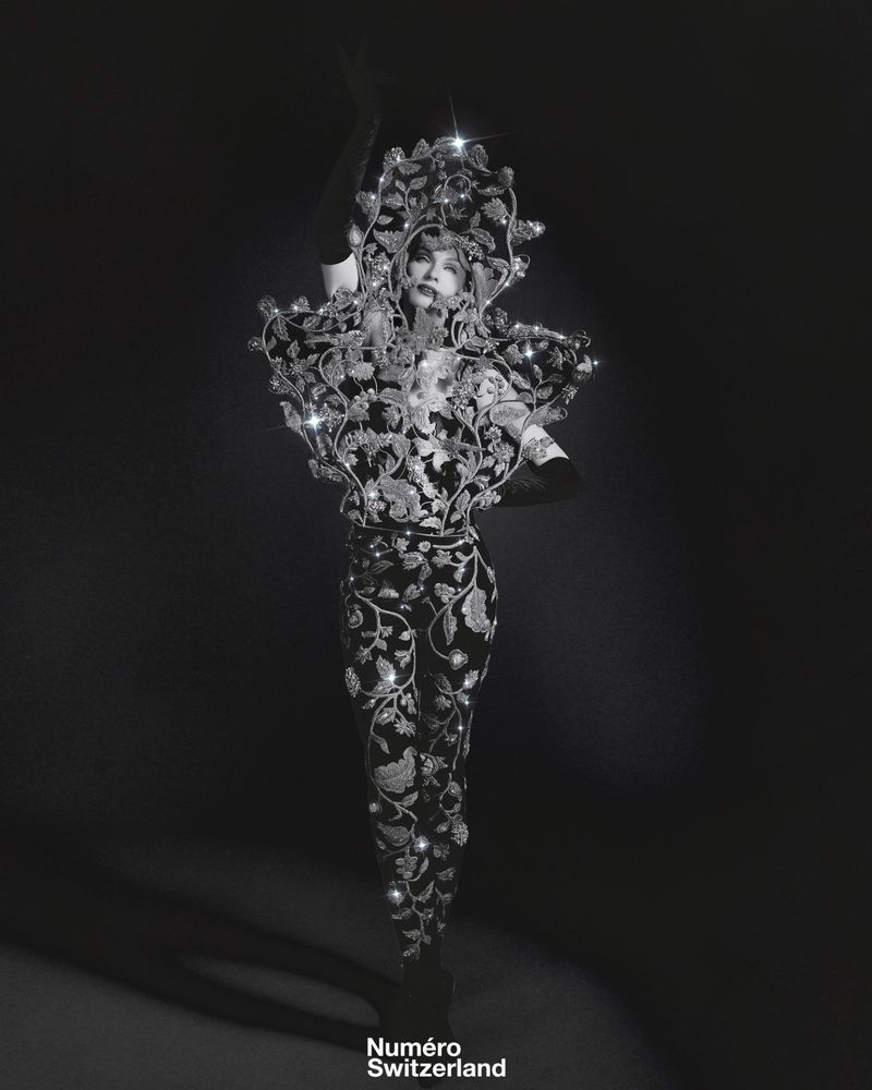 A person wears an elaborate, dark floral couture garment with embellishments in a studio, showcasing dramatic fashion and artistic design.