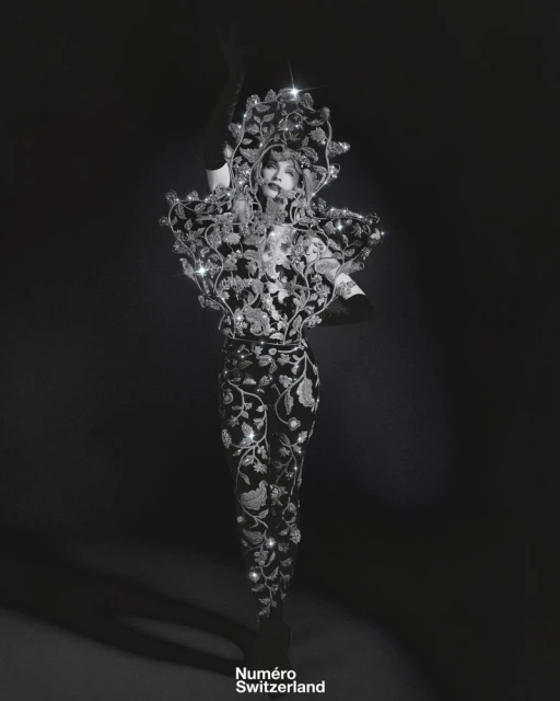 A person wears an elaborate, dark floral couture garment with embellishments in a studio, showcasing dramatic fashion and artistic design.