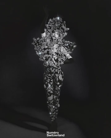 A person wears an elaborate, dark floral couture garment with embellishments in a studio, showcasing dramatic fashion and artistic design.