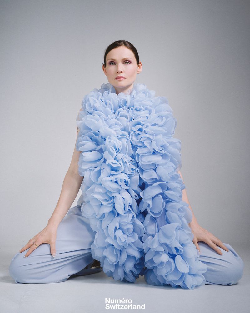 A woman in light blue pants and a matching ruffled shawl sitting against a plain grey background in a fashion photoshoot.