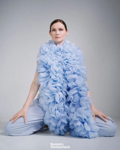 A woman in light blue pants and a matching ruffled shawl sitting against a plain grey background in a fashion photoshoot.
