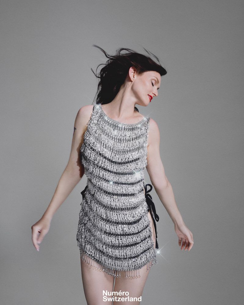 A young adult woman poses in a sleeveless silver and black beaded dress in a studio, against a neutral grey backdrop for fashion editorial.