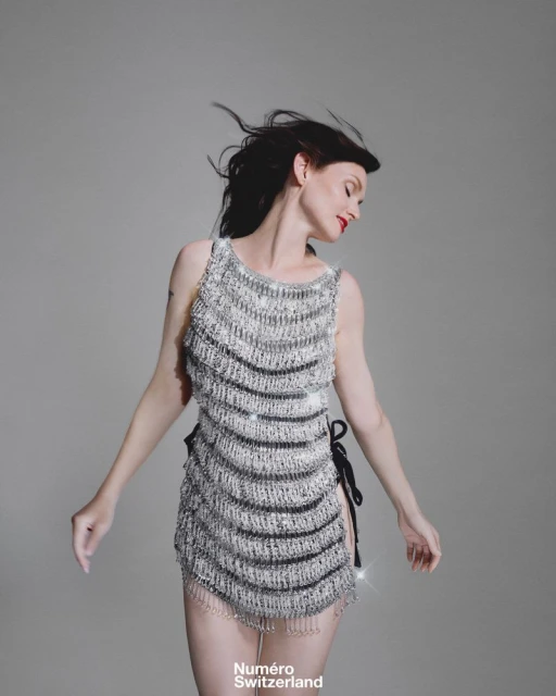 A young adult woman poses in a sleeveless silver and black beaded dress in a studio, against a neutral grey backdrop for fashion editorial.