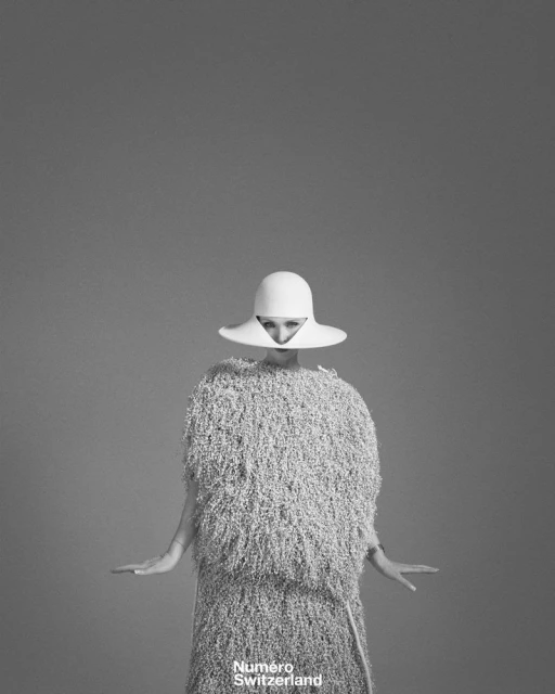 Monochrome fashion portrait of a woman in a textured dress and white hat against a gray backdrop. Fashion editorial style.