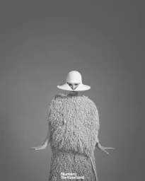 Model wearing a structured white hat and textured fringe dress against a grey studio background for Numéro Switzerland