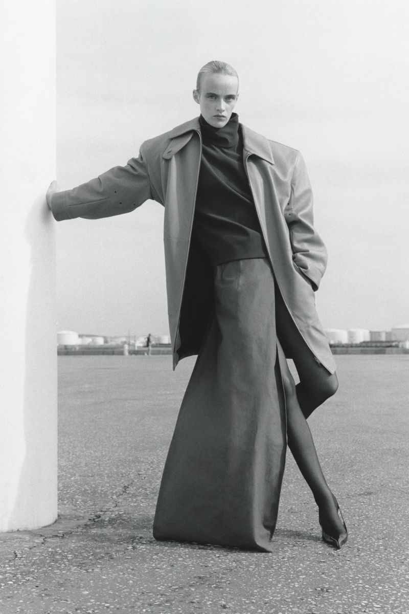 Fashion shot of adult woman wearing oversized trench coat, long skirt, heels, and black tights, leaning against a column outdoors.