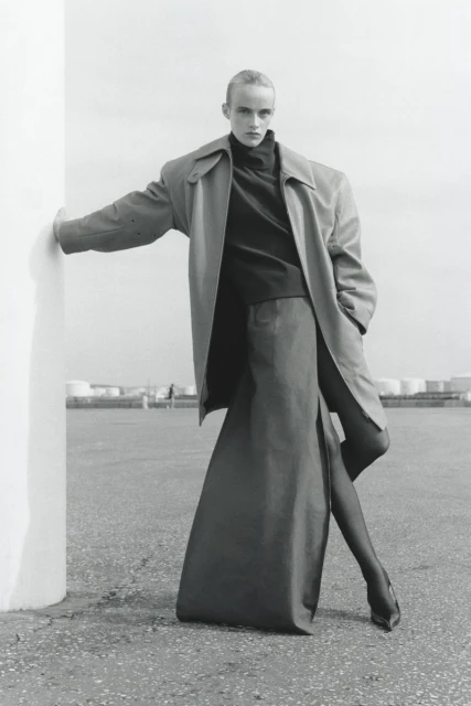 Fashion shot of adult woman wearing oversized trench coat, long skirt, heels, and black tights, leaning against a column outdoors.