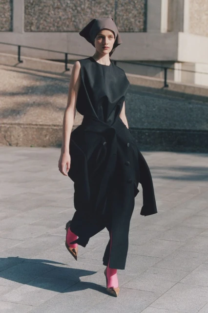 Woman in an oversized black dress and gray headscarf strides confidently on a concrete surface in bright sunlight with building in background.