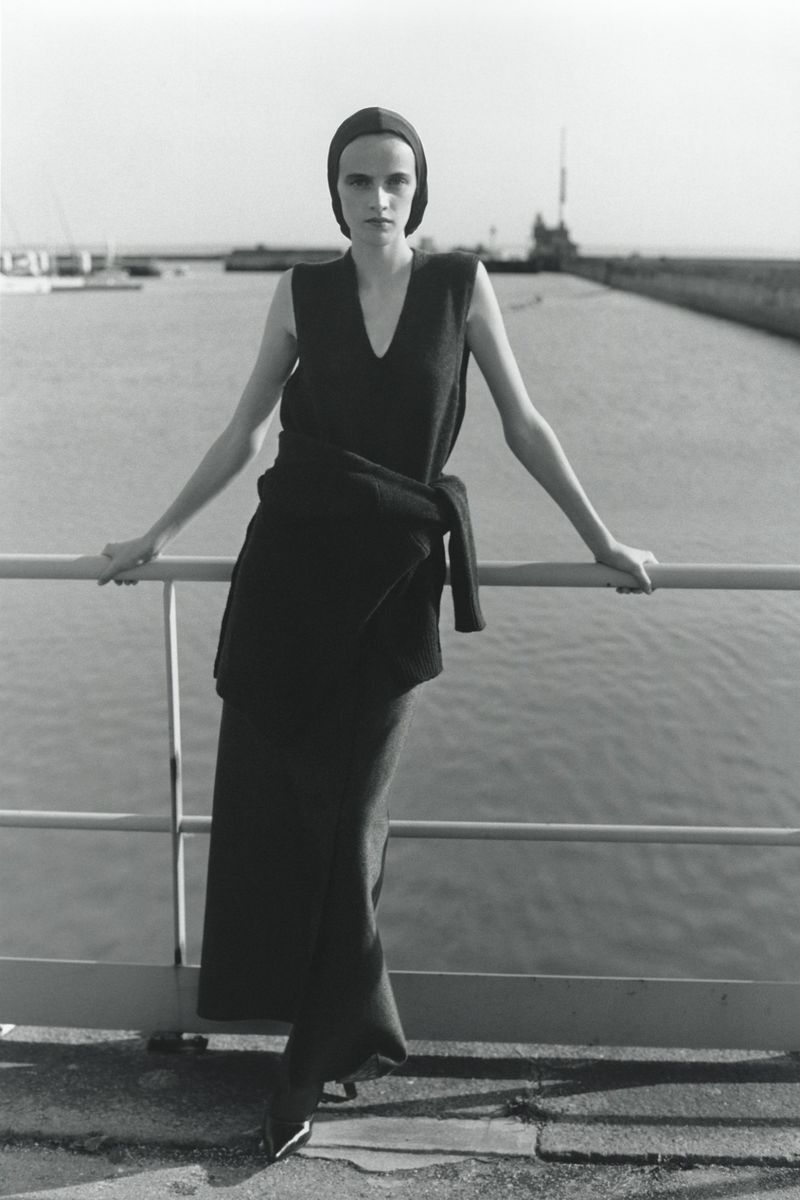 A slender adult woman poses on a bridge in an elegant black sleeveless dress against a waterfront background on a sunny day.