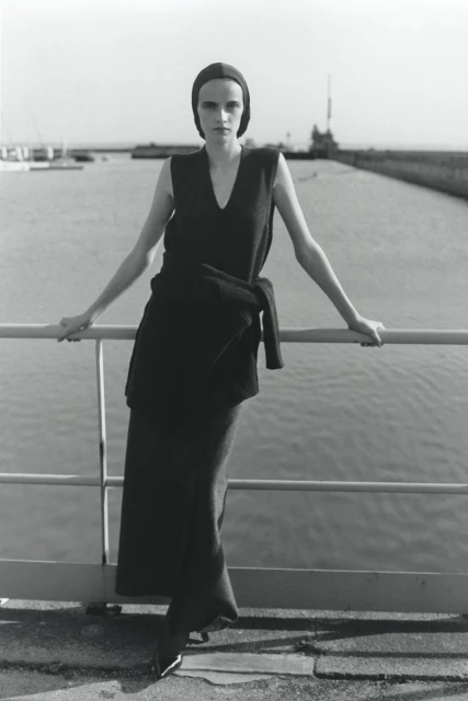 A slender adult woman poses on a bridge in an elegant black sleeveless dress against a waterfront background on a sunny day.