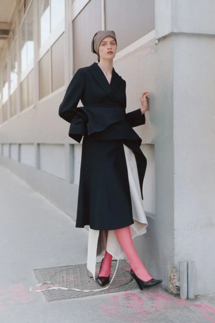 Elegant woman in a black peplum blazer and skirt with pink tights, posing against an urban wall in natural light fashion shot.
