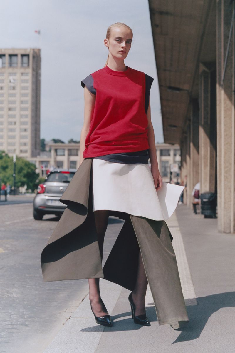 A slender fashion model in a red top and draped skirt poses on a city street near a building and car for an outdoor editorial shot.
