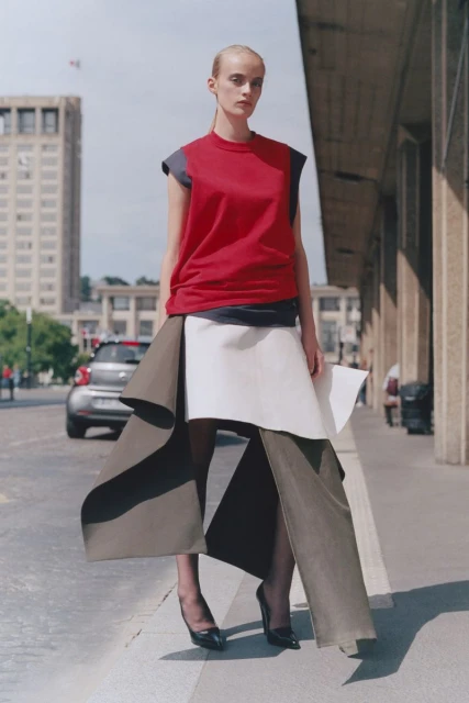 A slender fashion model in a red top and draped skirt poses on a city street near a building and car for an outdoor editorial shot.