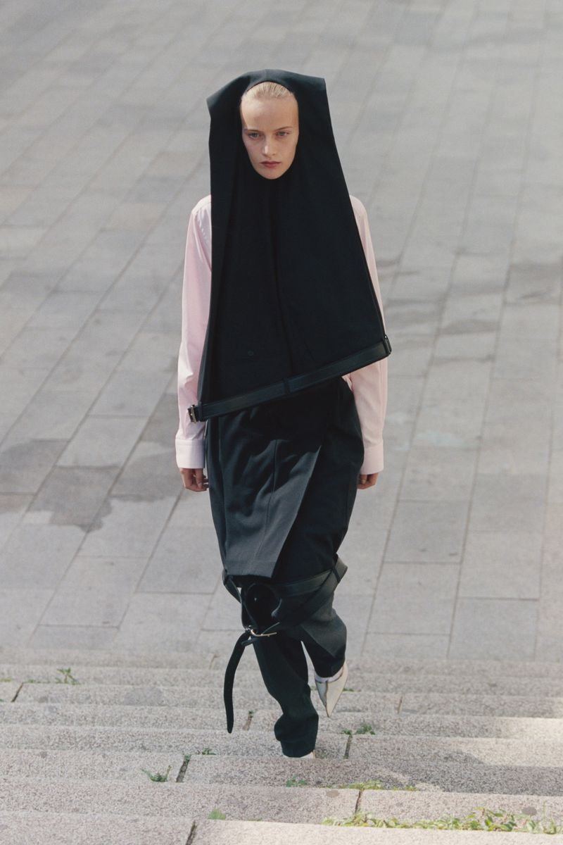 High-angle view of a fashion model in black avant-garde hood and pants with a pink shirt descending concrete steps outdoors.