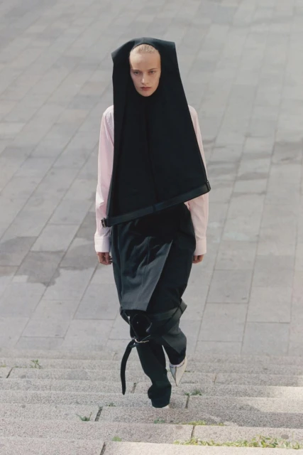 High-angle view of a fashion model in black avant-garde hood and pants with a pink shirt descending concrete steps outdoors.