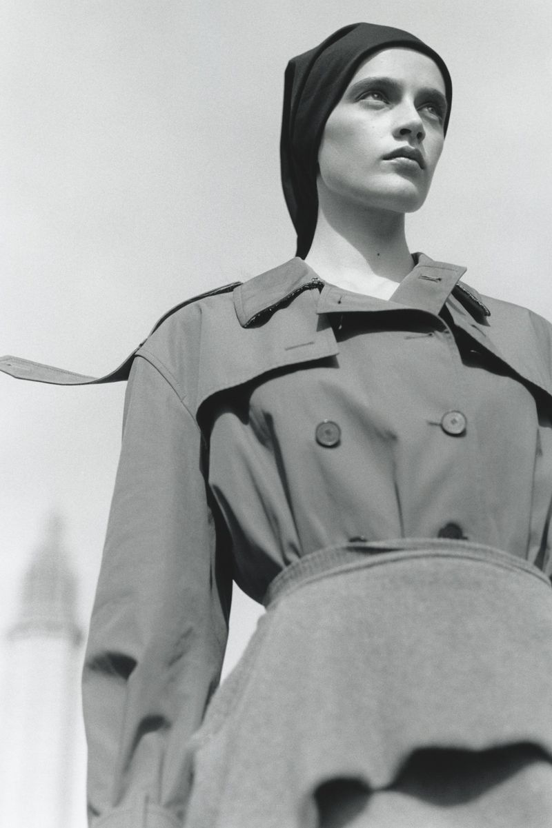 Monochrome fashion portrait of a model wearing a headscarf and belted trench coat against a clear sky background
