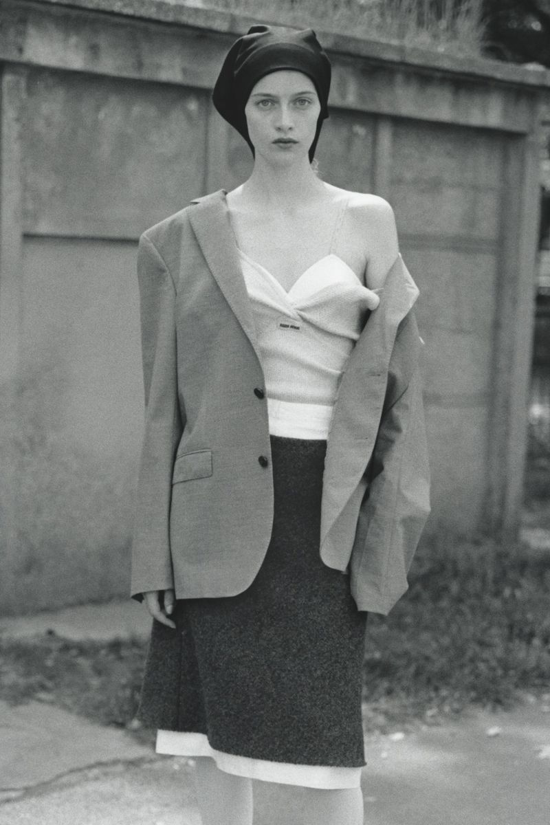 Model wearing dark headscarf, white layered top, dark blazer and skirt against a concrete wall in a black and white editorial photo.