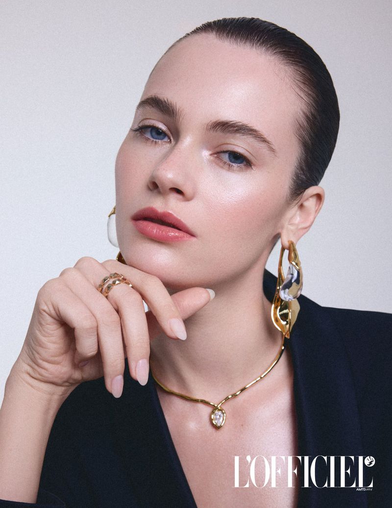 Headshot of a fair-skinned woman with blue eyes, wearing jewelry and a black blazer against a light background. Beauty and fashion.