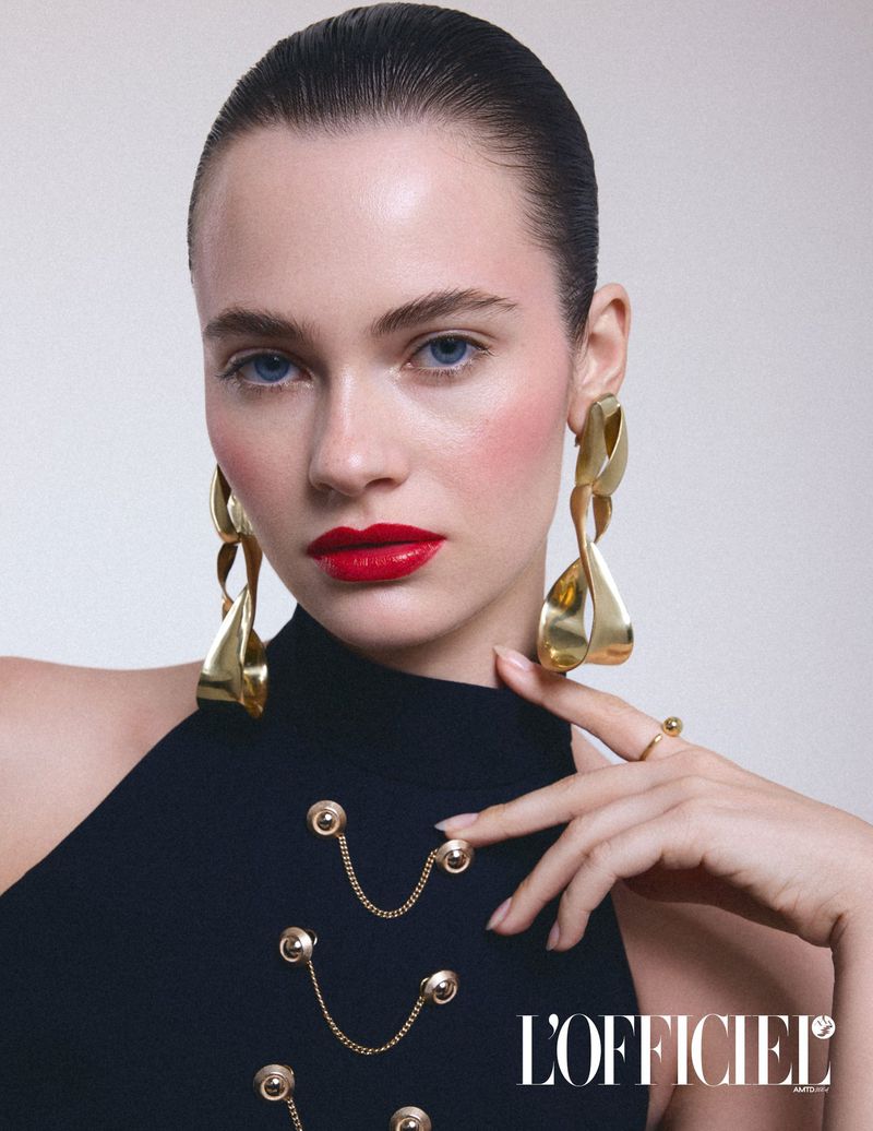 Close-up beauty portrait of a young woman with red lipstick wearing gold earrings and a black top with gold button details in a photo studio.