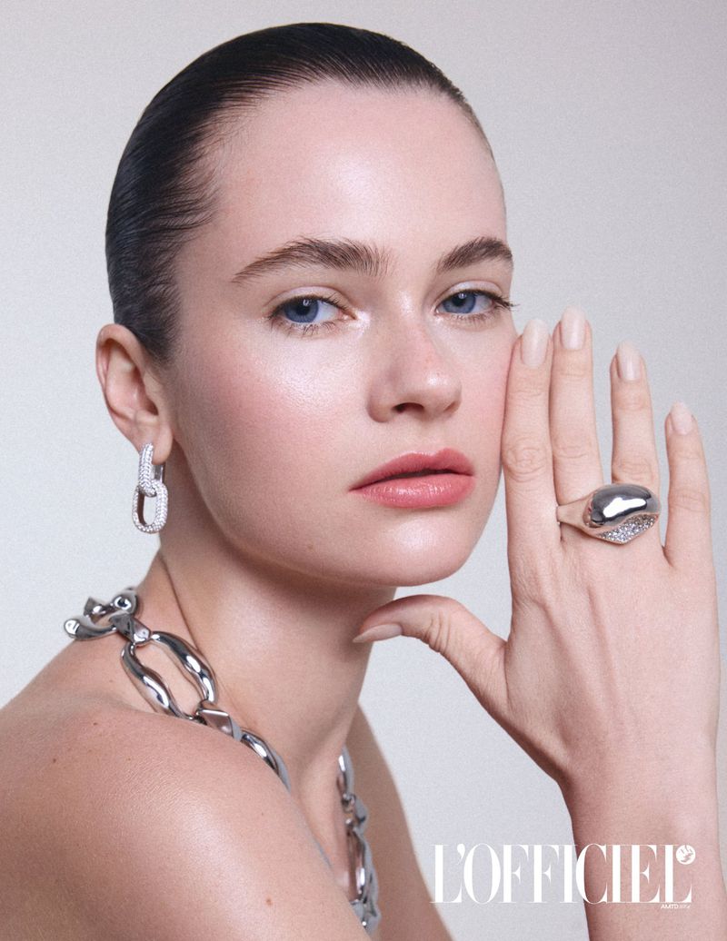 Close-up portrait of a young adult woman with silver jewelry including earrings, a ring, and a chain necklace against a white backdrop.