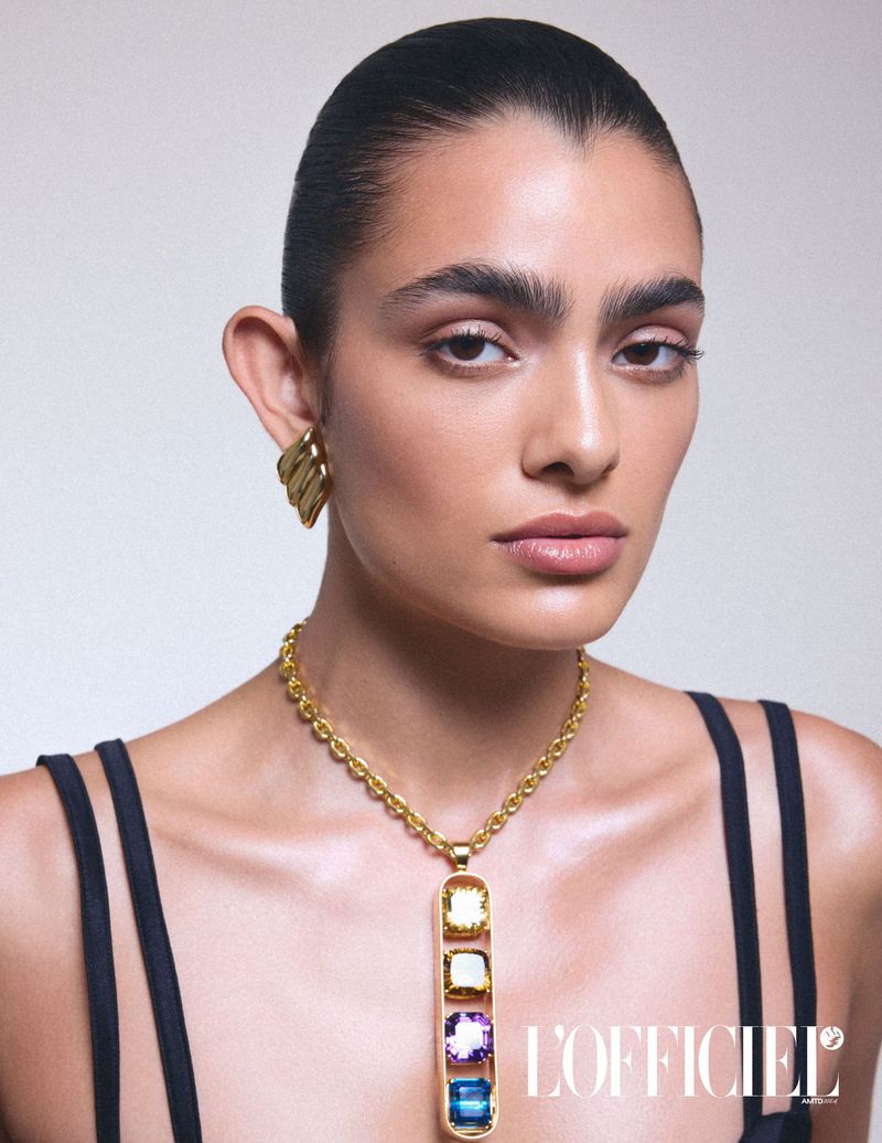 Close-up studio shot of a woman with dark hair and golden jewelry, including earrings and a necklace with colored gemstones.