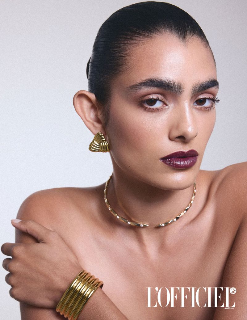 Close-up studio shot of a woman modeling gold jewelry, including earrings, choker necklace, and a bracelet, against a light background.