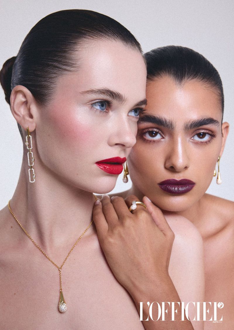 Two adult women with gold jewelry pose together in a studio shot with professional lighting, showcasing dark lipstick and a fashion magazine logo.