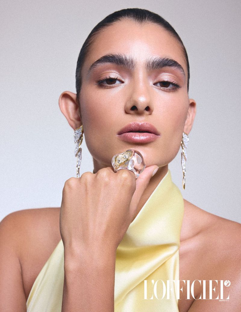Headshot of a model with tan skin, wearing ornate jewelry, makeup, and a light yellow dress in front of a light grey background.