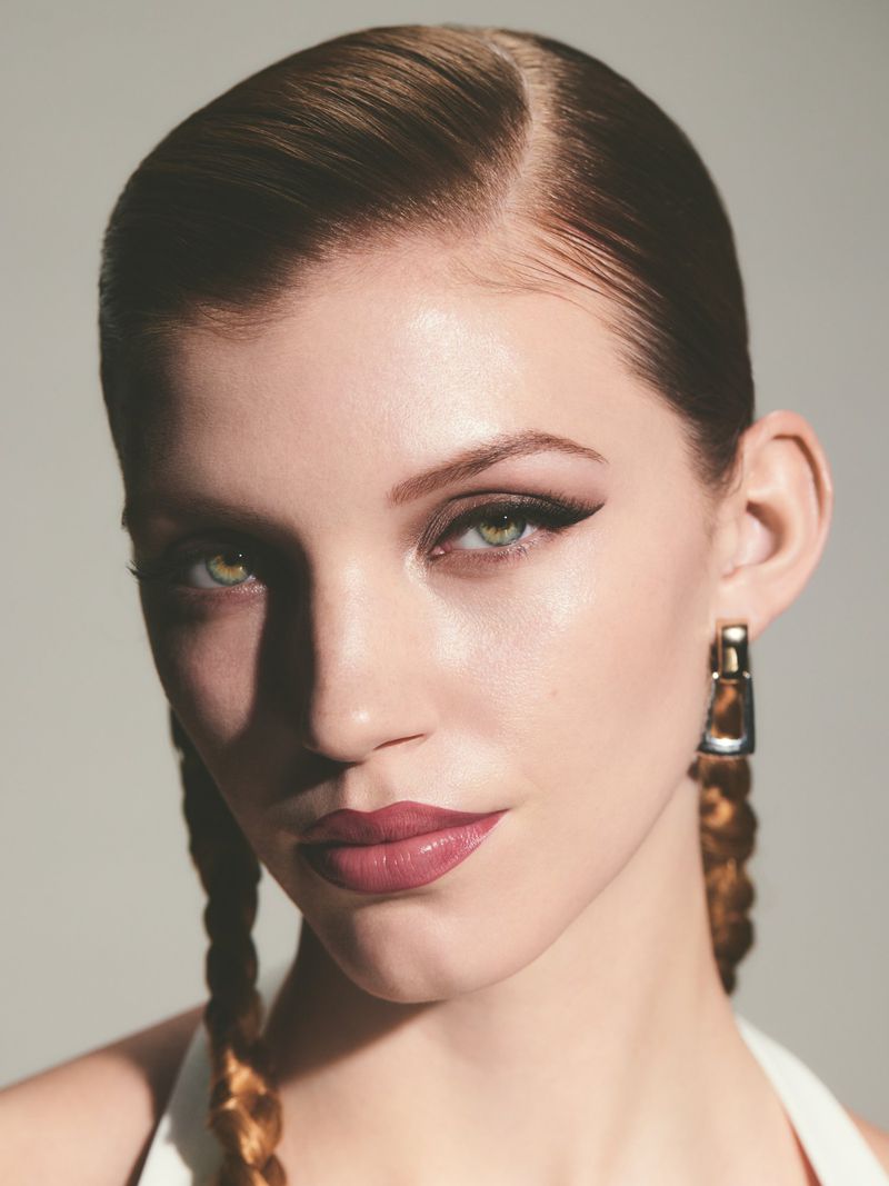 Close-up portrait of a young adult woman with green eyes and braided hair, wearing eye makeup and lipstick, against a plain background.
