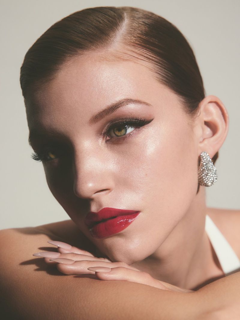 Close-up studio shot of a stylish adult woman with glossy red lips, elegant updo, and sparkly earrings, posing with hands under her chin.