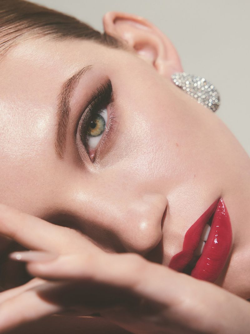 Close-up of a young adult female face with glossy red lipstick, hand near her face, jewelry earring, and dramatic eye makeup.