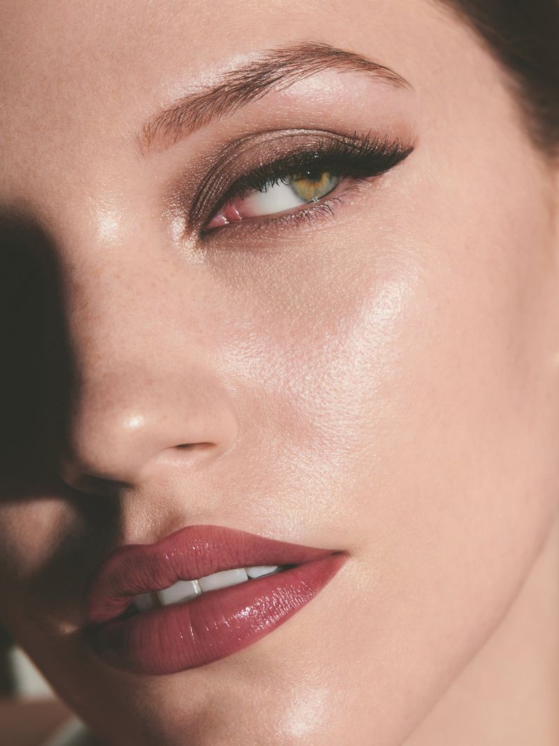 Close-up portrait of a woman's face, showcasing glossy red lips, defined eyeliner, and soft, natural makeup. Professional beauty shot.