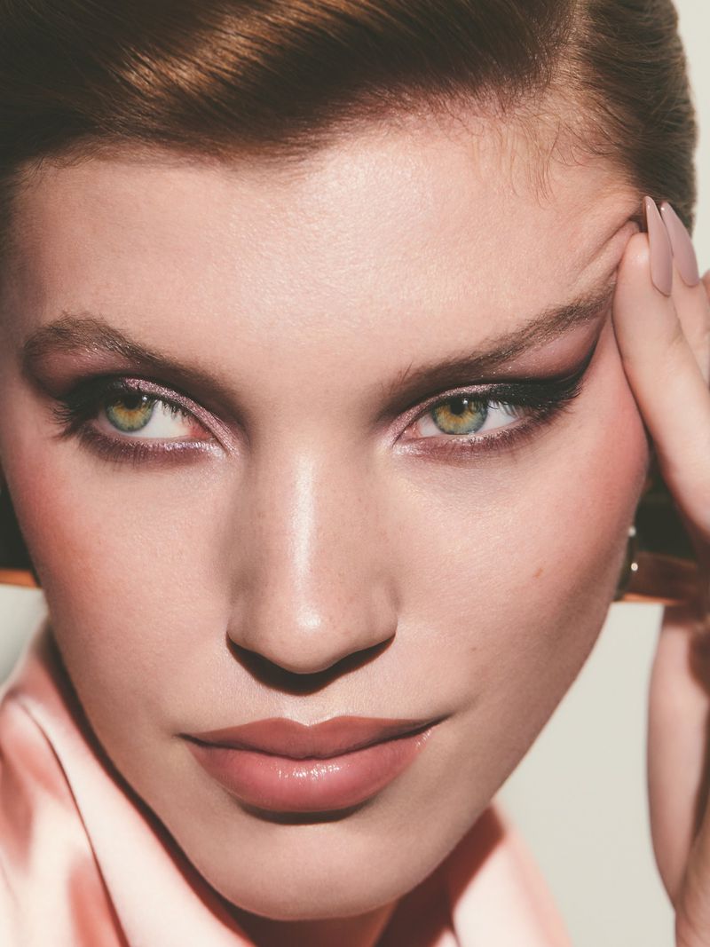 Close-up portrait of a woman with fair skin, green eyes, wearing eye makeup and pink lipstick, touching her forehead with her fingers.