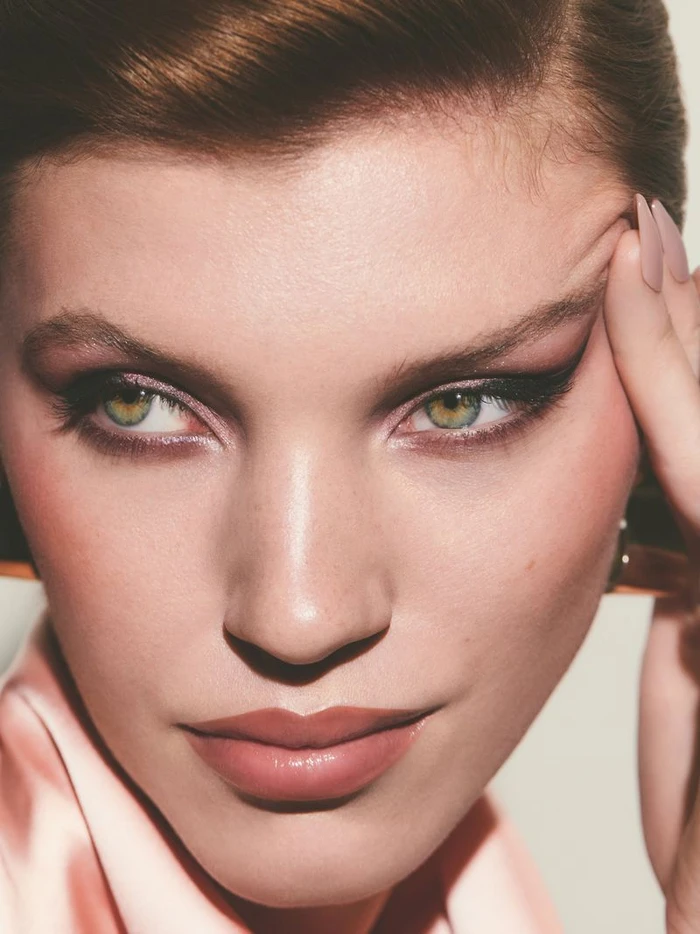 Close-up beauty portrait of a woman with green eyes, winged eyeliner, and pink makeup resting her hand on her temple.
