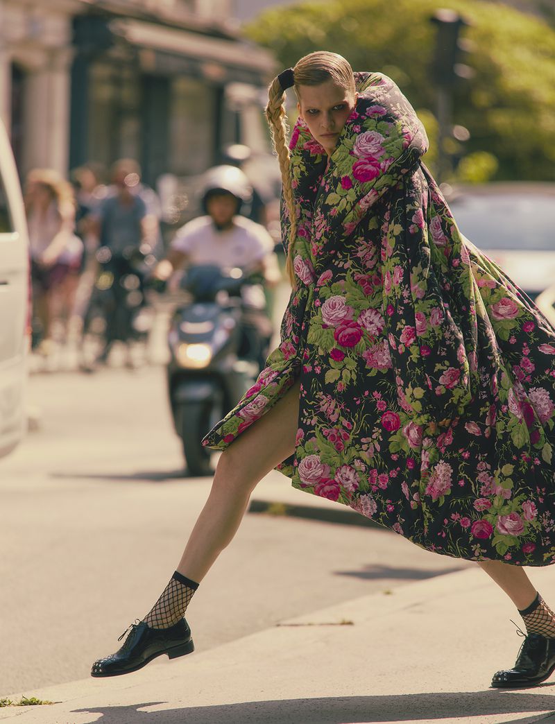 An adult female strides confidently on a city street in a bold floral coat, fishnet socks, and black shoes, with blurred urban background.