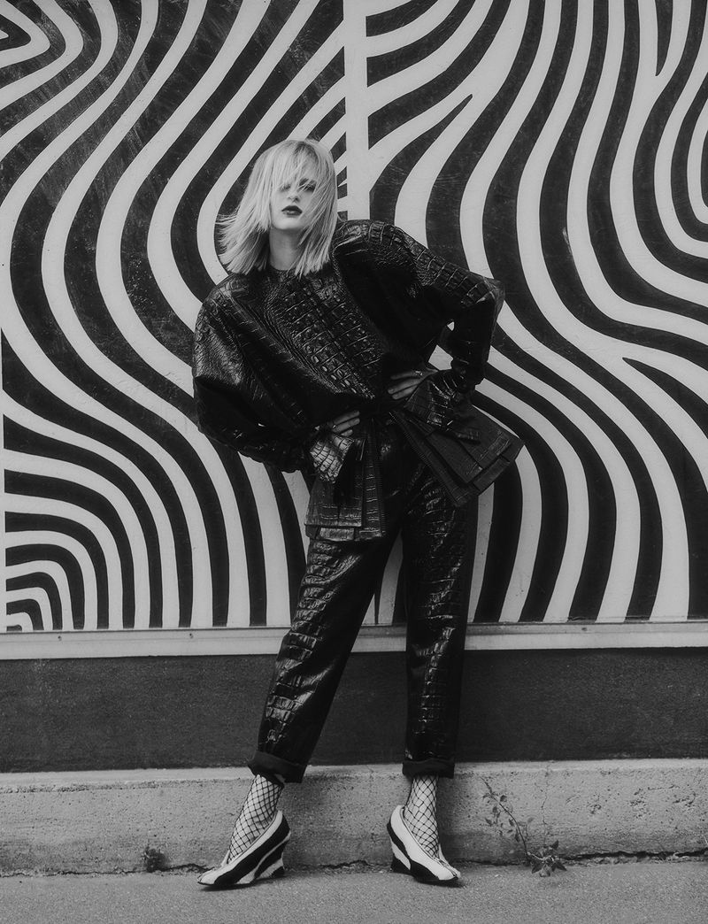A woman poses in a black outfit against a black and white patterned wall, showcasing fashion in an urban setting with unique wedge heels.