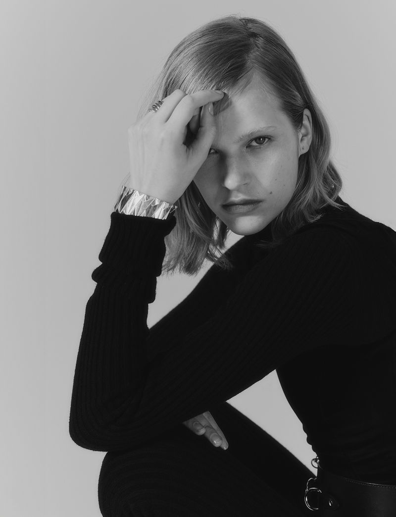 Black and white portrait of a woman in a black turtleneck with an ornate silver bracelet, sitting in front of a plain grey background.