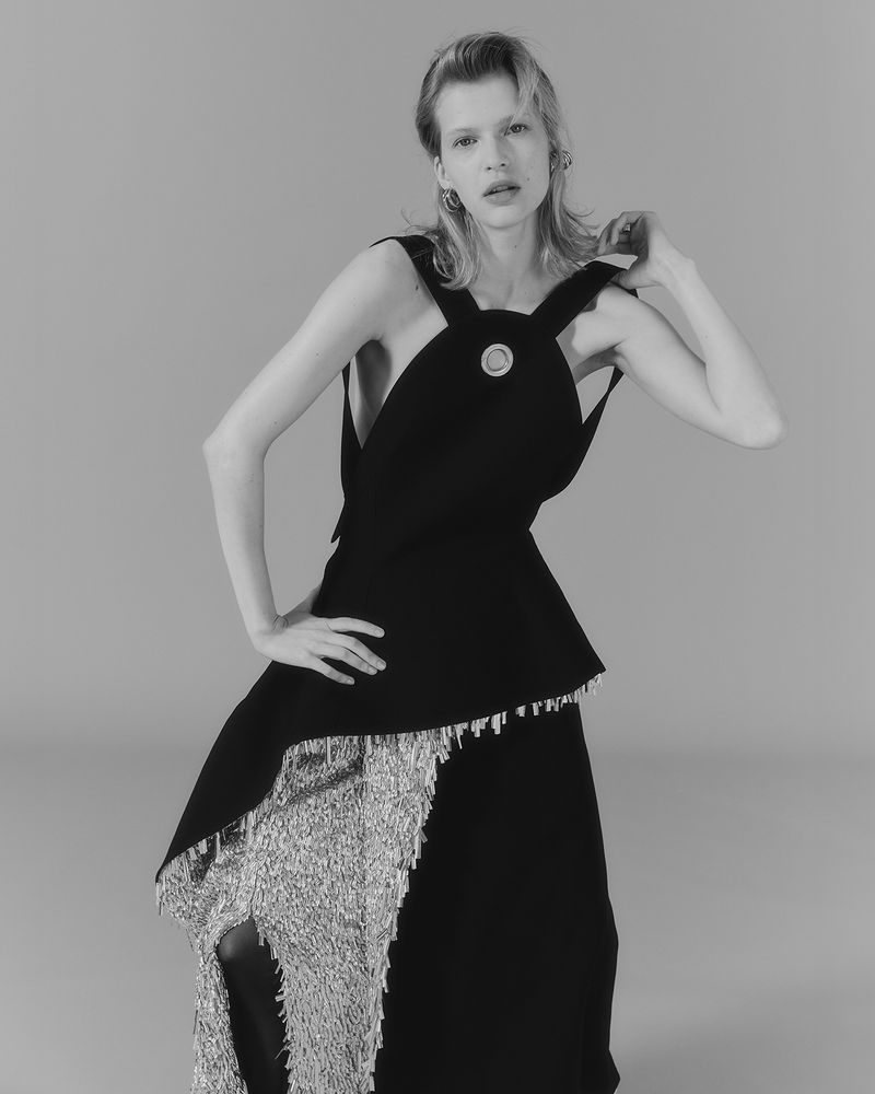 Monochrome portrait of a woman in a black top and silver fringe skirt, posing indoors with a neutral background, fashion-forward style.