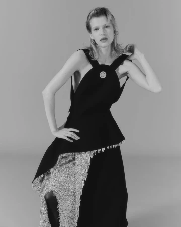 Monochrome portrait of a woman in a black top and silver fringe skirt, posing indoors with a neutral background, fashion-forward style.