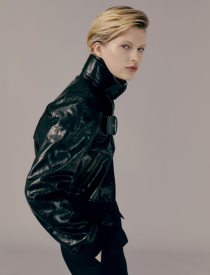 Blonde-haired woman poses in a shiny black leather jacket in a studio shot with a soft gray background and medium-shot framing.