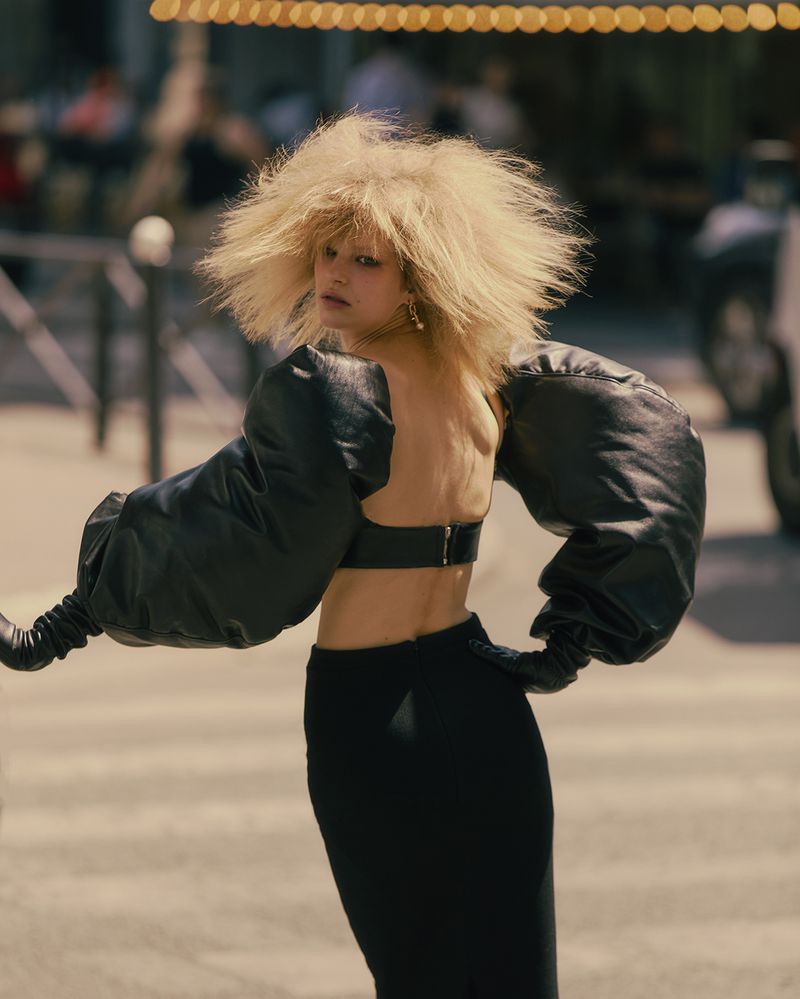 Fashionable adult woman with voluminous blonde hair poses in an open-back top and black skirt on a city street during daylight.