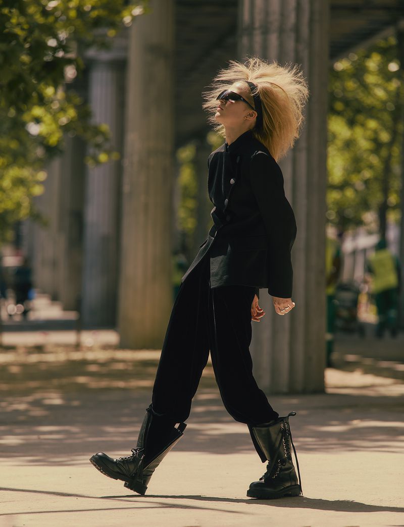 Fashionable adult woman with oversized blonde hair strides confidently outdoors in black jacket, black trousers, and tall black boots.